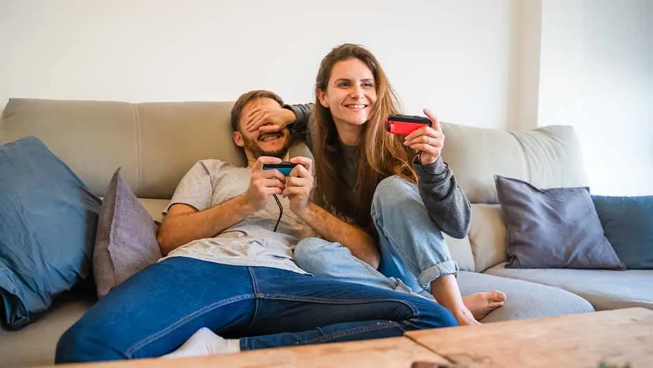 Een man en een vrouw zitten op een bank en spelen videospelletjes. De vrouw houdt de ogen van de man bedekt.
