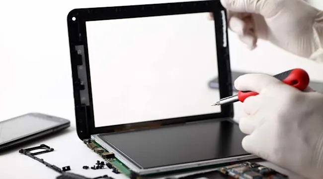 Hands with gloves hold a screwdriver to repair a tablet. Nearby are a phone, and electronic components on a white surface.