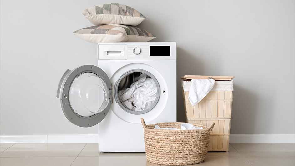 A laundry room scene: A white washing machine with an open door, two pillows on top, and laundry baskets.