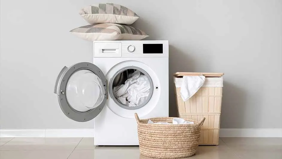 A laundry room scene: A white washing machine with an open door, two pillows on top, and laundry baskets.