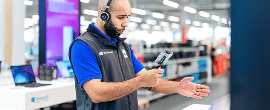Een man met een koptelefoon en een blauw shirt met een grijs vest houdt een telefoon vast in een winkel.