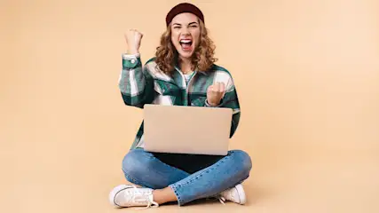 Vrouw zit gekruist met een laptop, draagt een geruite jas en muts, en balt haar vuisten in een gebaar van vreugde tegen een beige achtergrond.