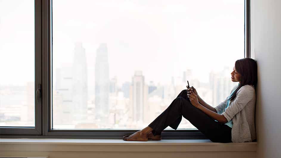 Een vrouw zit op een vensterbank met een telefoon in haar handen, kijkend naar buiten door een groot raam met uitzicht op gebouwen.