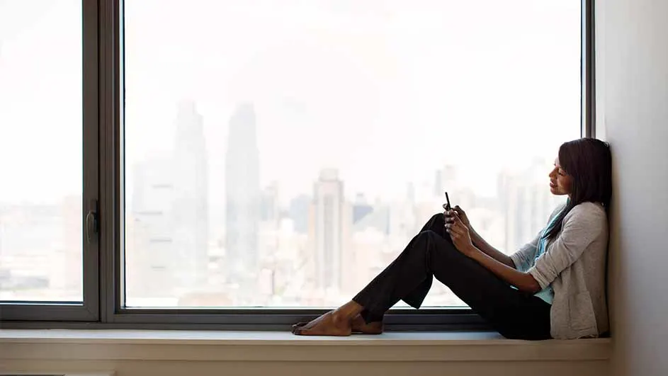 Een vrouw zit op een vensterbank met een telefoon in haar handen, kijkend naar buiten door een groot raam met uitzicht op gebouwen.