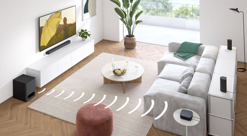 Lichte woonkamer met wit wandmeubel, zwarte soundbar en aparte subwoofer, grijze hoekbank, rond salontafeltje, vloerlamp en grote planten bij hoge ramen.