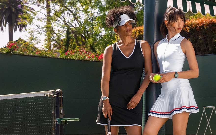 Twee tennisspeelsters in stijlvolle jurken, een zwart en een wit, poseren naast het net, elk met een elegante smartwatch om de pols.