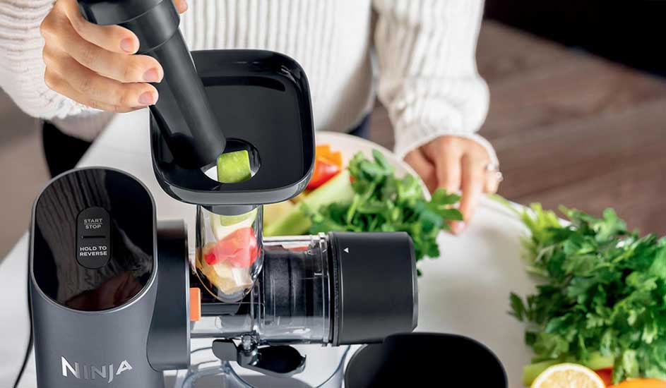 Op de afbeelding stopt iemand stukjes groente in een zwarte Ninja slowjuicer, omringd door verse peterselie en citroen op een witte tafel.