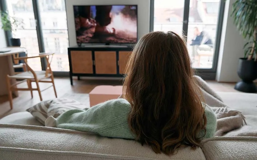 Een vrouw zit op een bank in een moderne woonkamer, kijkt ontspannen naar een televisie die een spannende scène toont.