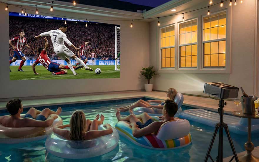 Mensen drijven in opblaasbanden in een binnenzwembad terwijl een projector een voetbalmatch op een grote muur toont.