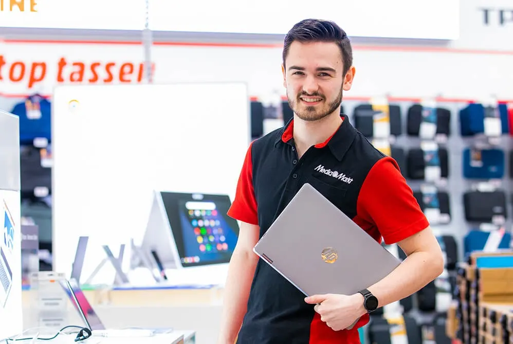 Een man in een rood-zwart shirt met een grijze laptop in een elektronicawinkel. Op de achtergrond staan schappen met producten.