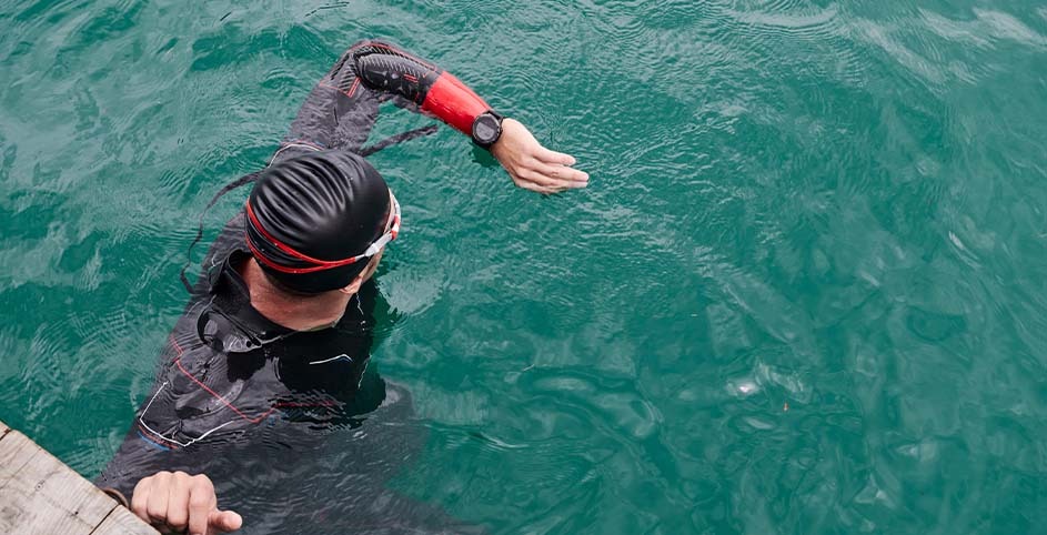 A swimmer in a black cap and wetsuit is in green water near a wooden dock, with a watch visible on their extended arm.