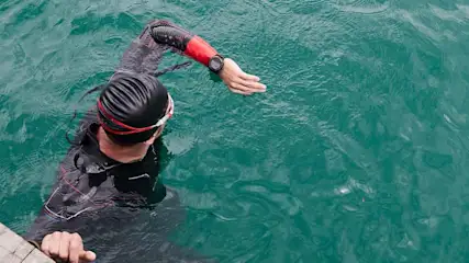 A swimmer in a black cap and wetsuit is in green water near a wooden dock, with a watch visible on their extended arm.