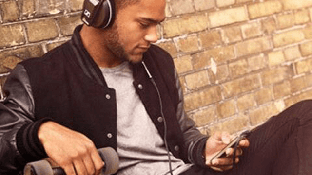 Man zit met koptelefoon, jas en T-shirt tegen een bakstenen muur, met een telefoon en gewichten.