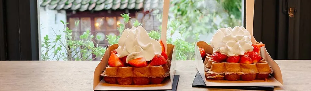Twee porties Belgische wafels geserveerd in kartonnen bakjes, belegd met verse aardbeien en een toef slagroom, gepresenteerd op een vensterbank met uitzicht op een groene tuin en traditioneel dak.