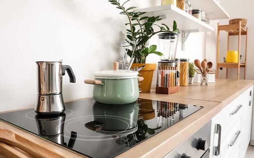 A modern kitchen with a glass hob, moka pot, green saucepan, spices, and plants on wooden worktops; no visible brand name appears.
