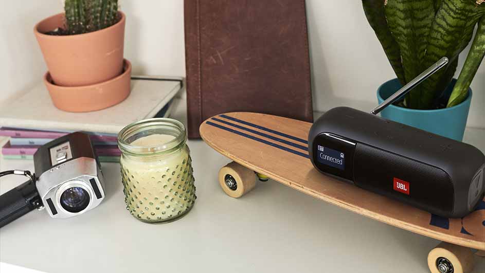 Een camera, kaars, boek, JBL-speaker op een skateboard, en planten op een witte tafel.