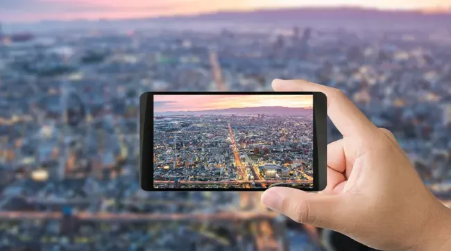 Een hand houdt een zwarte smartphone vast met een foto van een stad bij zonsondergang. Op de achtergrond is een stad te zien.