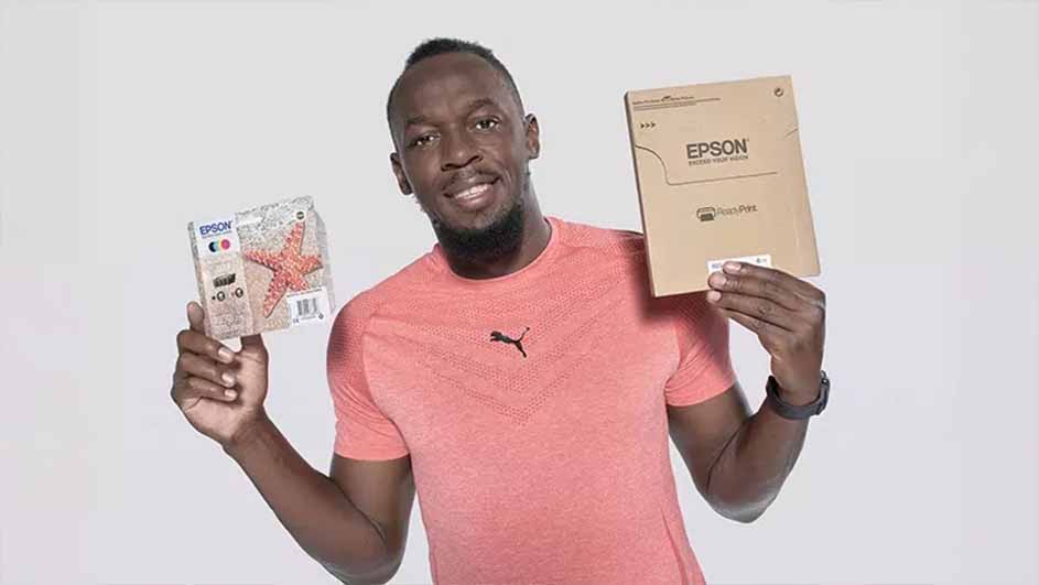 Een man met een roze shirt houdt een Epson verpakking en een verpakking met een zeester vast tegen een witte achtergrond.