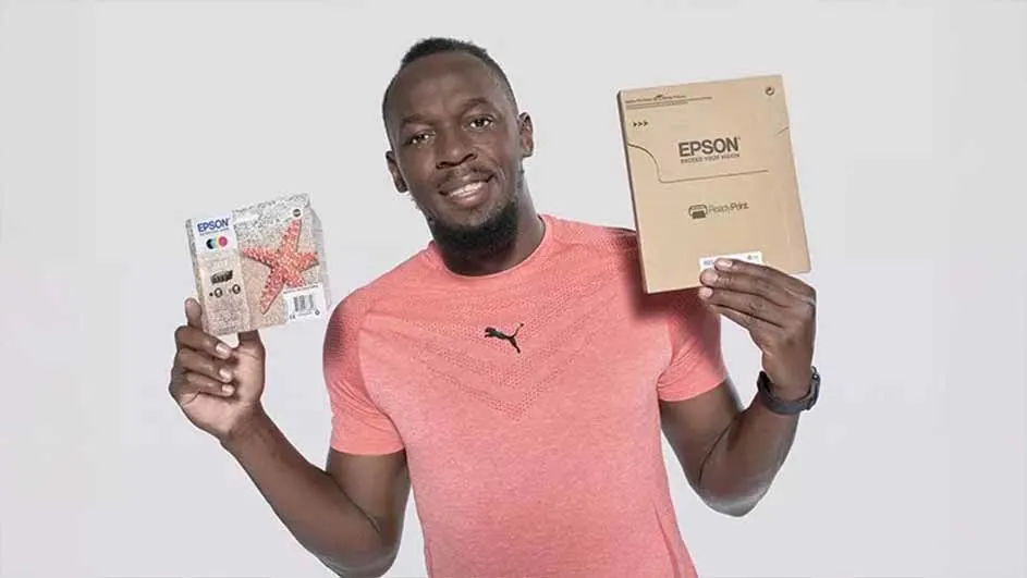 Een man met een roze shirt houdt een Epson verpakking en een verpakking met een zeester vast tegen een witte achtergrond.