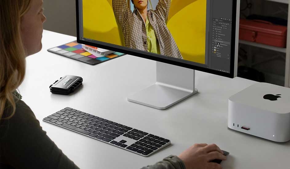 Person editing a bright yellow fashion image on a large monitor, with compact white desktop box and slim keyboard on desk.