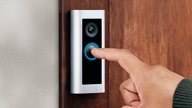 Een vinger drukt op een zilverkleurige Ring-videodeurbel met een camera boven een blauw verlichte knop op een houten deur.