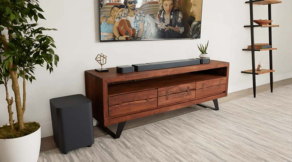 Een woonkamer met een houten tv-meubel, een grijze subwoofer en een tv aan de muur. Een plant en een open kast staan naast het meubel.
