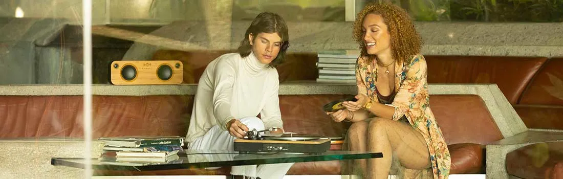 Two people relax on a low sofa by a glass table, choosing records and playing them on a turntable in a bright, leafy living room.