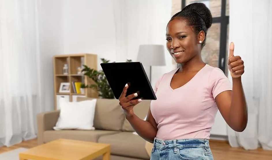 A woman in a living room holds a tablet, smiles at the camera and gives a thumbs-up; sofa, shelves and a lamp behind her.