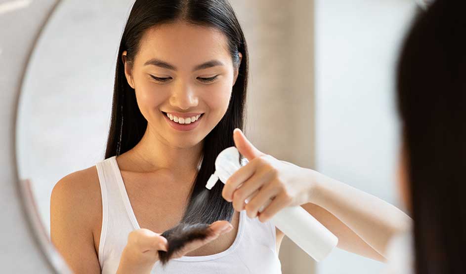 A smiling woman sprays product onto her long dark hair while looking into the mirror in soft, warm light.