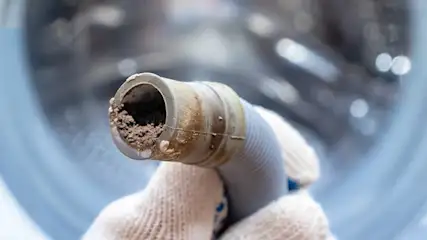 Een verstopte afvoerslang van een wasmachine wordt vastgehouden door een hand met een witte handschoen. De buis zit vol met vuilafzetting en kalkaanslag. Op de achtergrond is de trommel van de wasmachine zichtbaar.