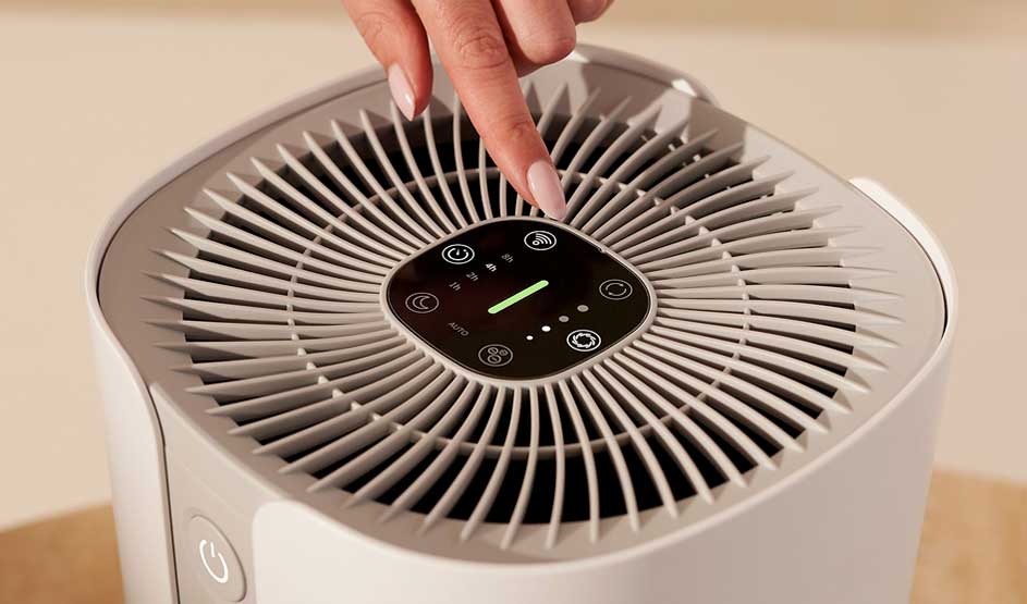 Hand pressing button on white air purifier with circular grille and digital touch control panel showing green indicator light
