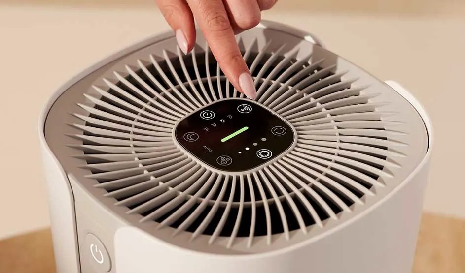 Hand pressing button on white air purifier with circular grille and digital touch control panel showing green indicator light
