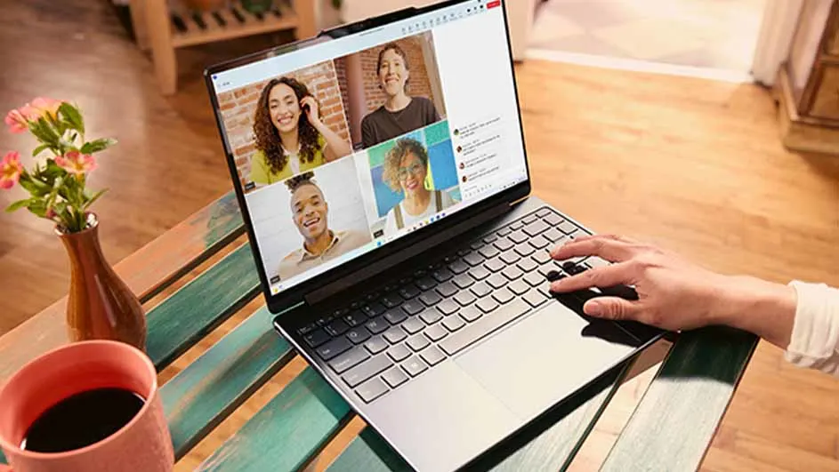 Een laptop op een tafel toont een videogesprek met vier personen. Een hand typt op het toetsenbord. Een kopje en vaas staan naast de laptop.