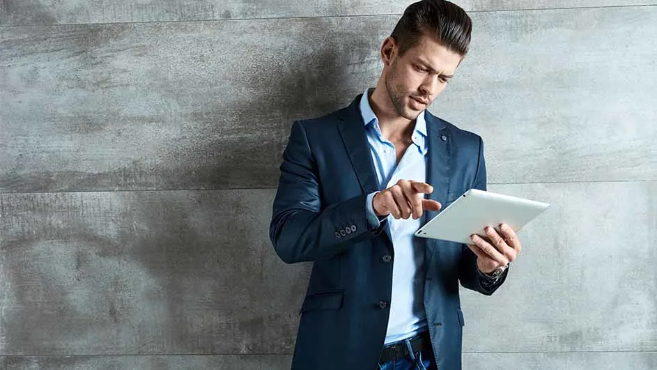 Een man met een blauw jasje en blauw overhemd gebruikt een tablet tegen een grijze achtergrond.