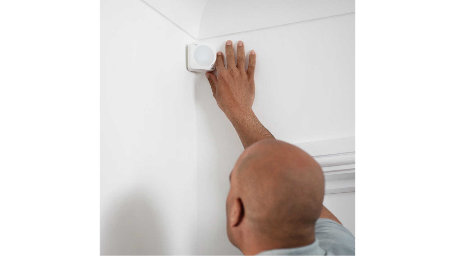 Een man bevestigt een wit, vierkant Ring-apparaat in een hoek van een witte muur, vlakbij een sierlijst.