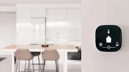 Zwarte thermostaat aan een witte muur, met een witte keuken en stoelen op de achtergrond.