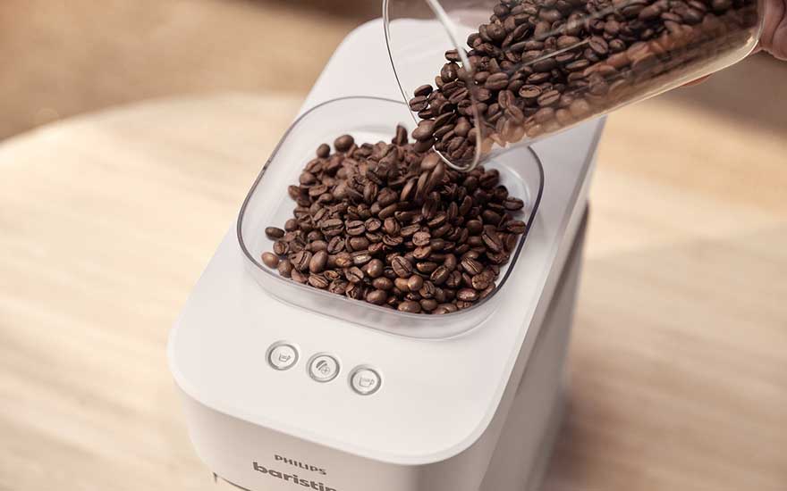 Dark coffee beans are poured into the hopper of a modern white coffee machine, standing neatly on a bright wooden surface.