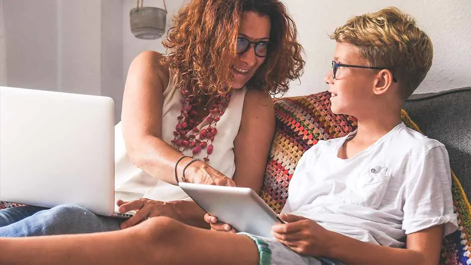 Een vrouw met een laptop en een jongen met een tablet zitten op een bank met een kleurrijk kussen.