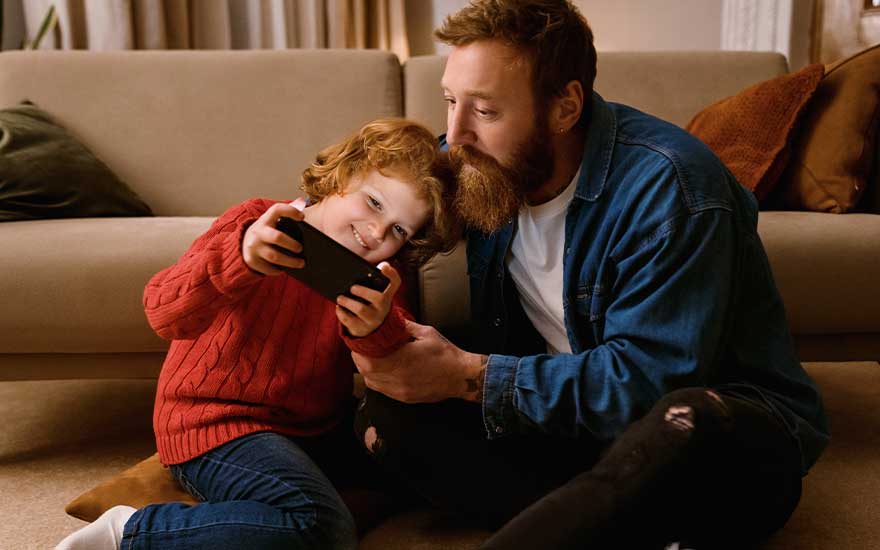 A child in a red jumper sits close to an adult on the floor as they happily watch something together on a smartphone screen.