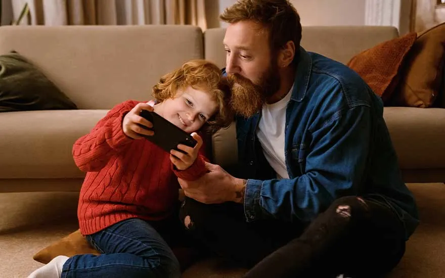 A child in a red jumper sits close to an adult on the floor as they happily watch something together on a smartphone screen.