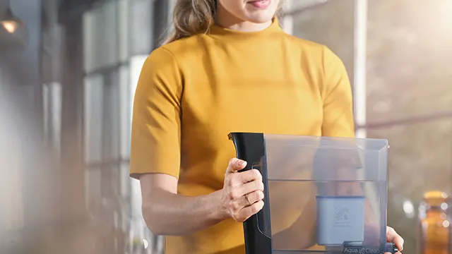 Een vrouw in een gele trui houdt een doorzichtige waterkan met een zwart handvat vast in een lichte kamer.