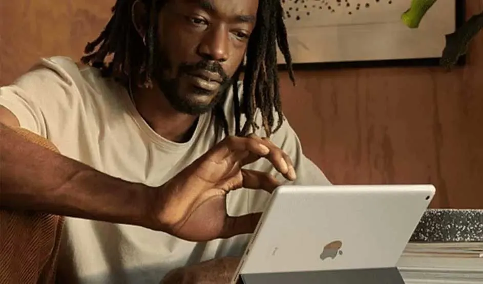 Een man zit binnen en gebruikt een iPad op een standaard; hij kijkt geconcentreerd en raakt het scherm aan, met warme verlichting en houtwand.