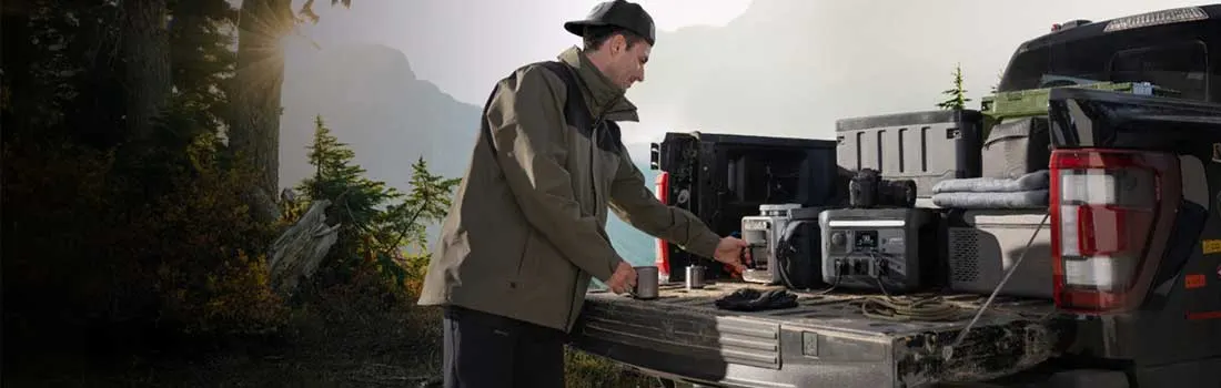 Een man bij een pick-up in de bergen gebruikt een draagbare stroomgenerator tussen kampeeruitrusting en koffers, omringd door natuur.