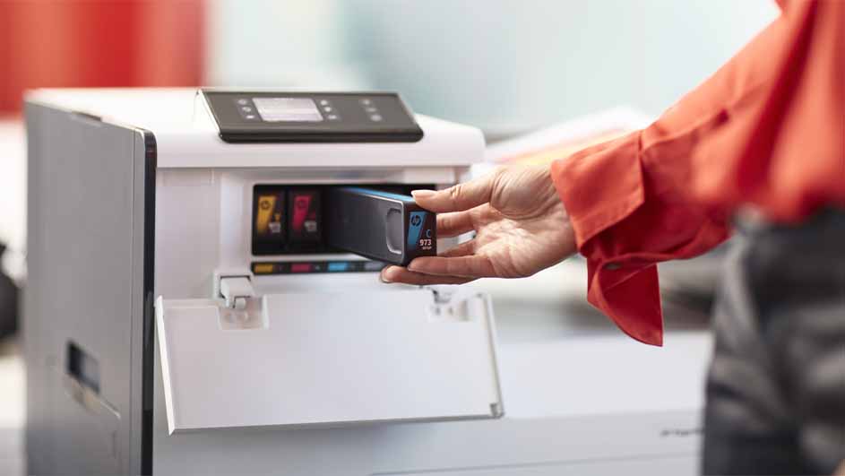 Een hand plaatst een zwarte inktcartridge in een witte printer met open klep. Kleuren zijn zichtbaar in de printer.