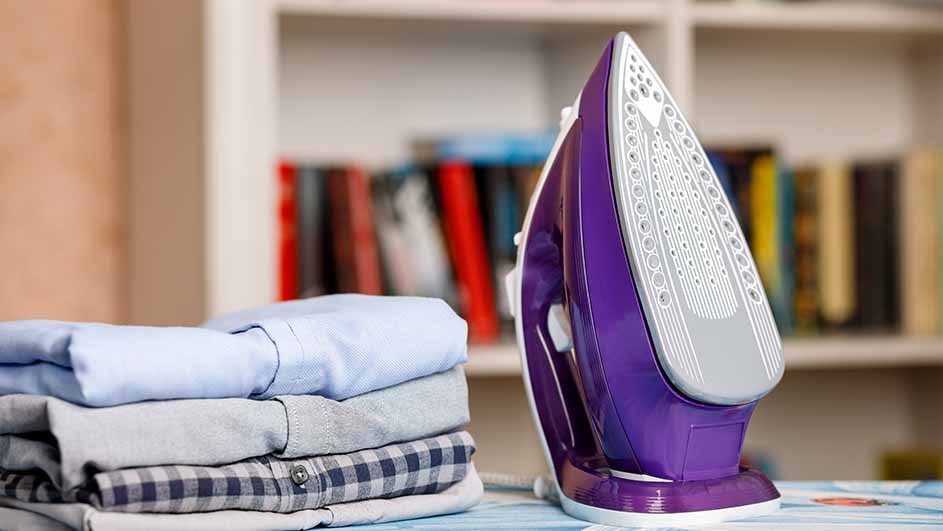 Een paars strijkijzer staat naast een stapel opgevouwen overhemden op een strijkplank, met een boekenkast op de achtergrond.