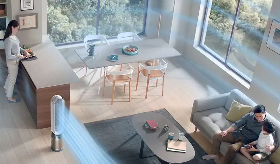 In a bright living room a tall air purifier blows cool air while one person cooks at the counter and a parent relaxes on the sofa with a child.