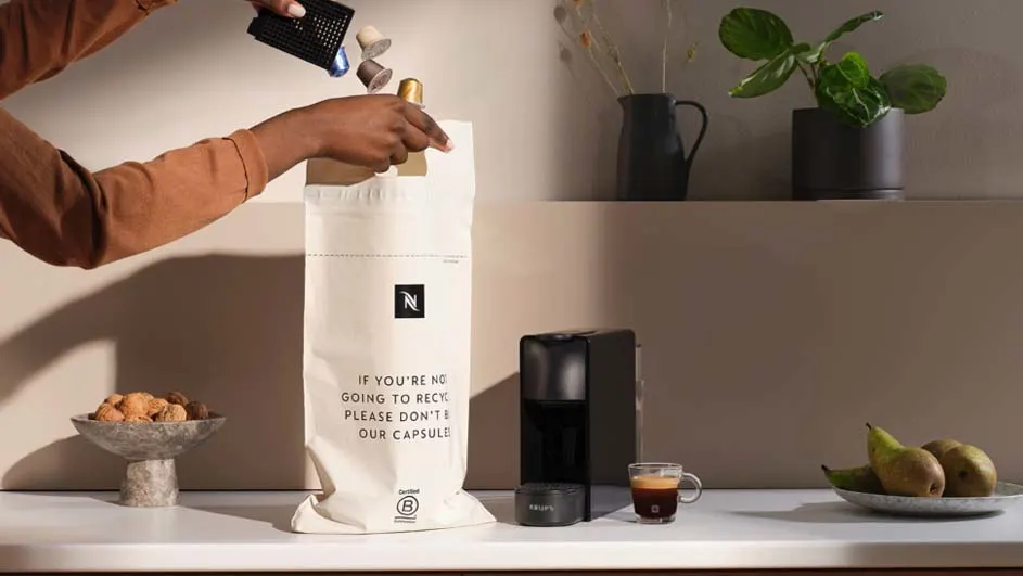 Iemand leegt koffiecapsules in een Nespresso recyclezak. Op het aanrecht staan een machine, koffie, fruit en koekjes.