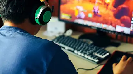 Een persoon met een blauw shirt en een groene koptelefoon speelt een computerspel met een toetsenbord en muis.