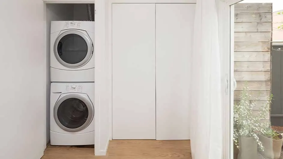 A laundry closet shows a stacked white washer and dryer next to folding white doors and a potted plant near a window.