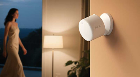 Een witte Aqara-sensor hangt aan de muur in een warme kamer, terwijl op de achtergrond een vrouw voorbijloopt.